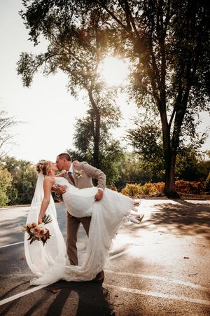 Groom dips bride in stunning white gown for a golden sunset kiss on a tree-lined road.