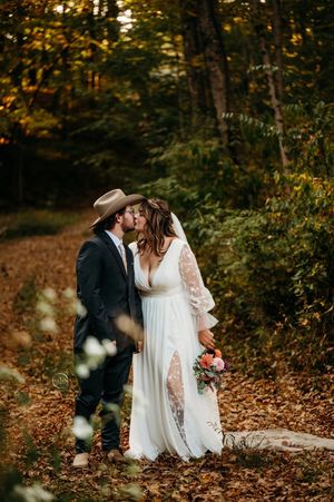 Bride and groom share a romantic autumn forest kiss, bride holding pink bouquet in flowing white gown.