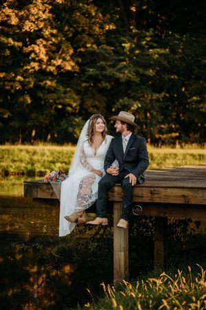 Bride and groom sit together on a wooden dock surrounded by lush autumn foliage at golden hour.