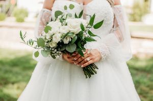Bride in lace wedding gown holds elegant white rose and eucalyptus bridal bouquet outdoors.