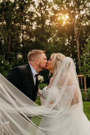 Bride and groom share a passionate kiss at sunset, bride's veil flowing dramatically in the breeze.