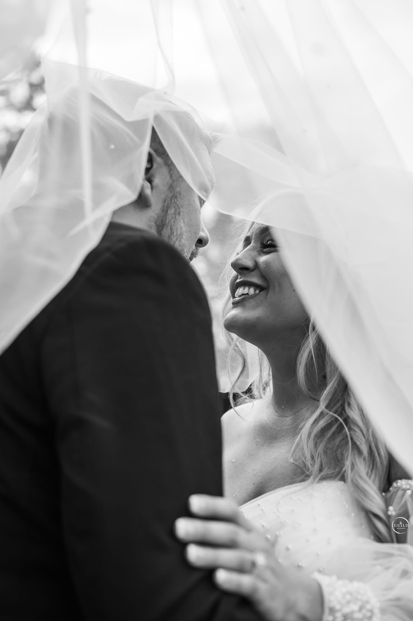 Black and white photo of smiling bride and groom sharing an intimate moment beneath her flowing veil.