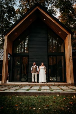 Bride and groom holding hands standing before a dramatic black A-frame cabin with large glass windows at dusk.