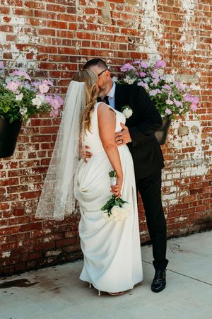A groom kisses his bride near a rustic brick wall adorned with hanging flower baskets during their wedding.