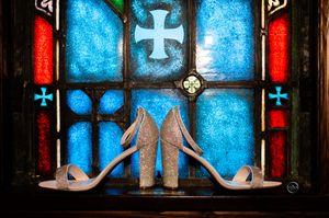 Glittery bridal heels displayed against colorful stained glass window with cross motifs.