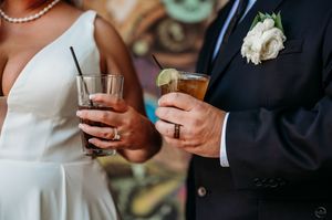 A bride and groom hold cocktail drinks during their wedding reception, showing their rings and wedding attire.