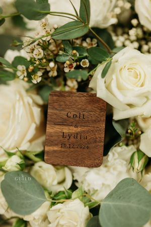 Wooden ring box engraved with Colt and Lydia 10.12.2024 nestled among white roses and eucalyptus greenery.