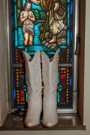 White cowboy boots displayed before a colorful stained glass church window depicting a baptism scene.
