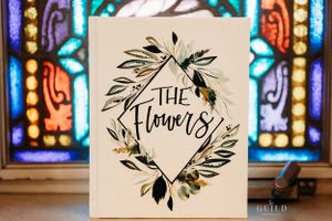 Decorative wedding book titled The Flowers with botanical design in front of stained glass.