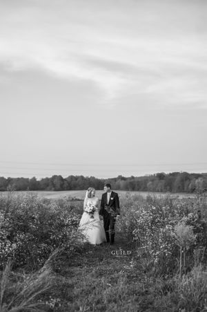 Black and white photo of bride and groom walking hand in hand through open field.