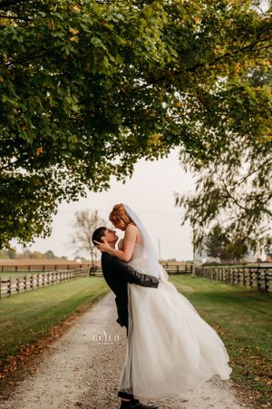 Groom lifting bride under lush green tree canopy on scenic farm pathway.
