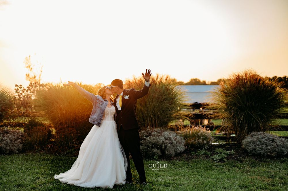 Bride and groom celebrating at golden sunset near a serene lake with raised hands.