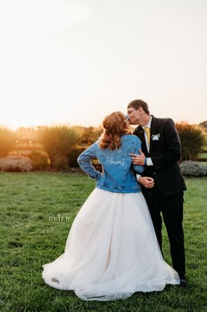 Bride in denim jacket kissing groom at golden sunset on green lawn.