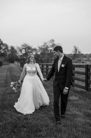 Black and white photo of smiling bride and groom holding hands along wooden fence.