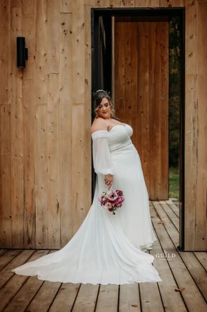 Bride in an off-shoulder white wedding gown with long sleeves holding a pink bouquet beside a wooden barn door.