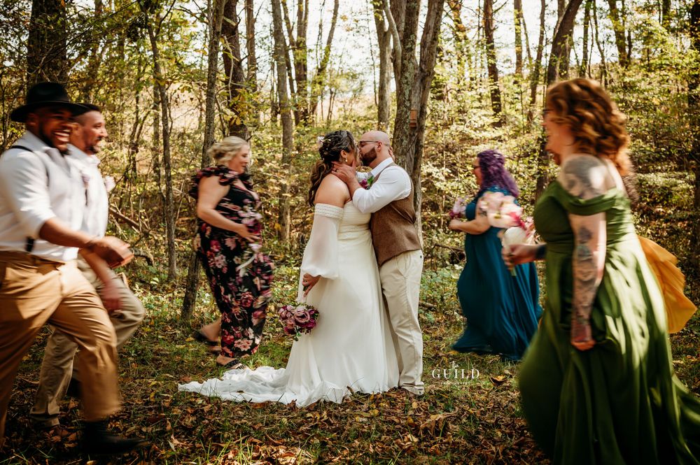 Bride and groom sharing a kiss during an intimate outdoor woodland wedding ceremony with wedding party nearby.