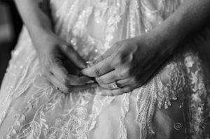Black and white close-up of bride's hands adjusting lace wedding gown with engagement ring visible.