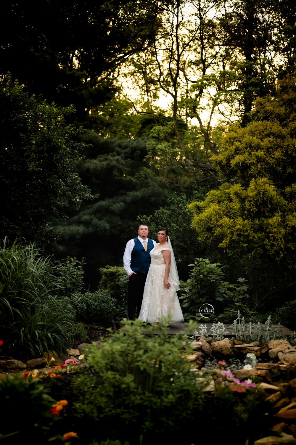 Bride and groom posing in lush garden at golden hour sunset surrounded by greenery.