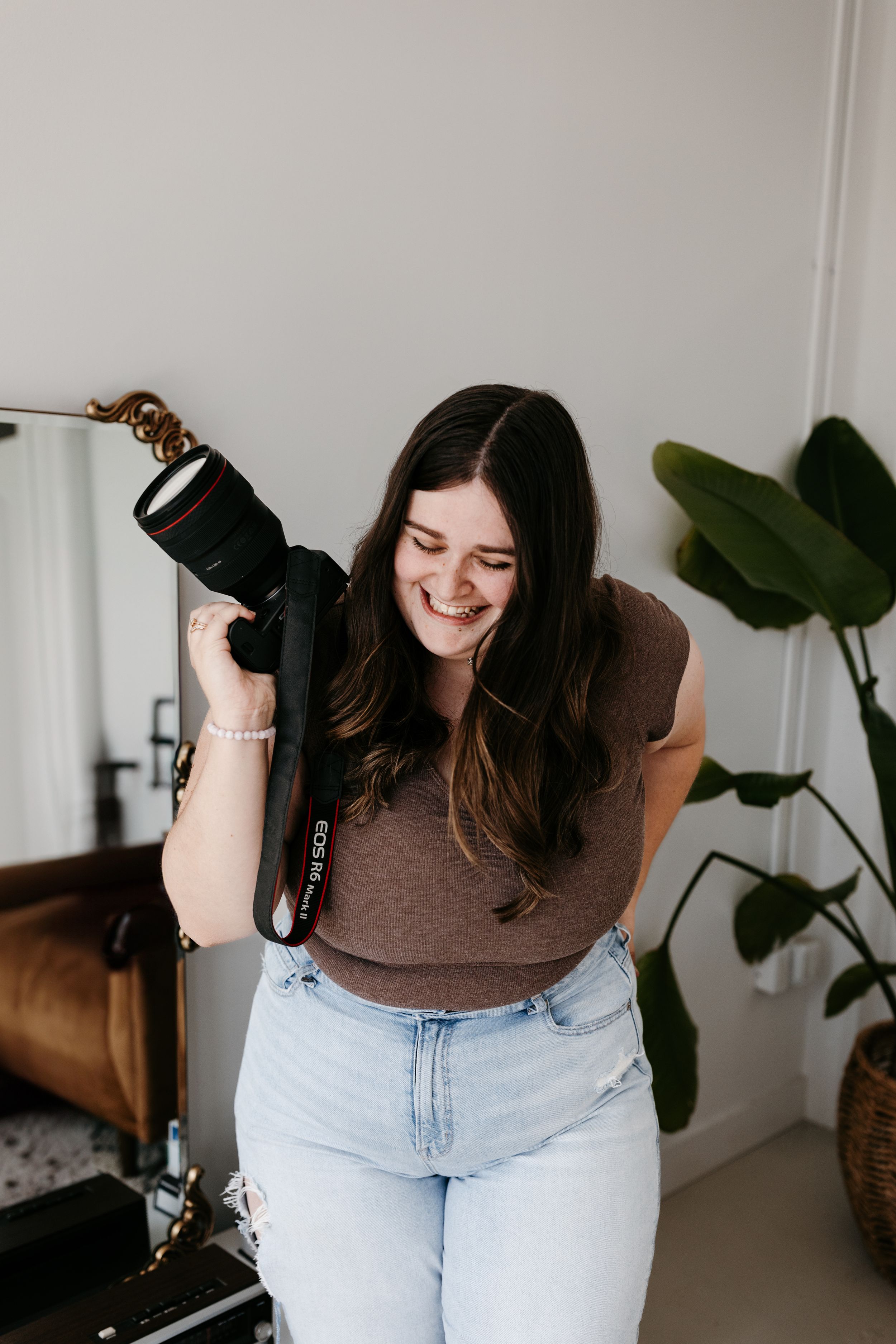 A photographer laughs while holding a camera in a cozy home setting with plants.
