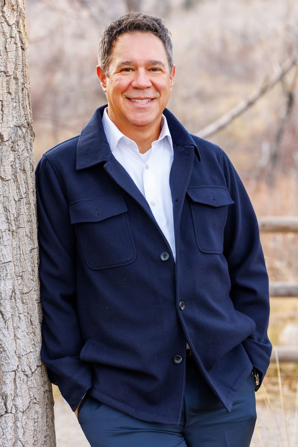 Professional portrait of someone wearing a navy peacoat and white dress shirt posing against a natural outdoor background.