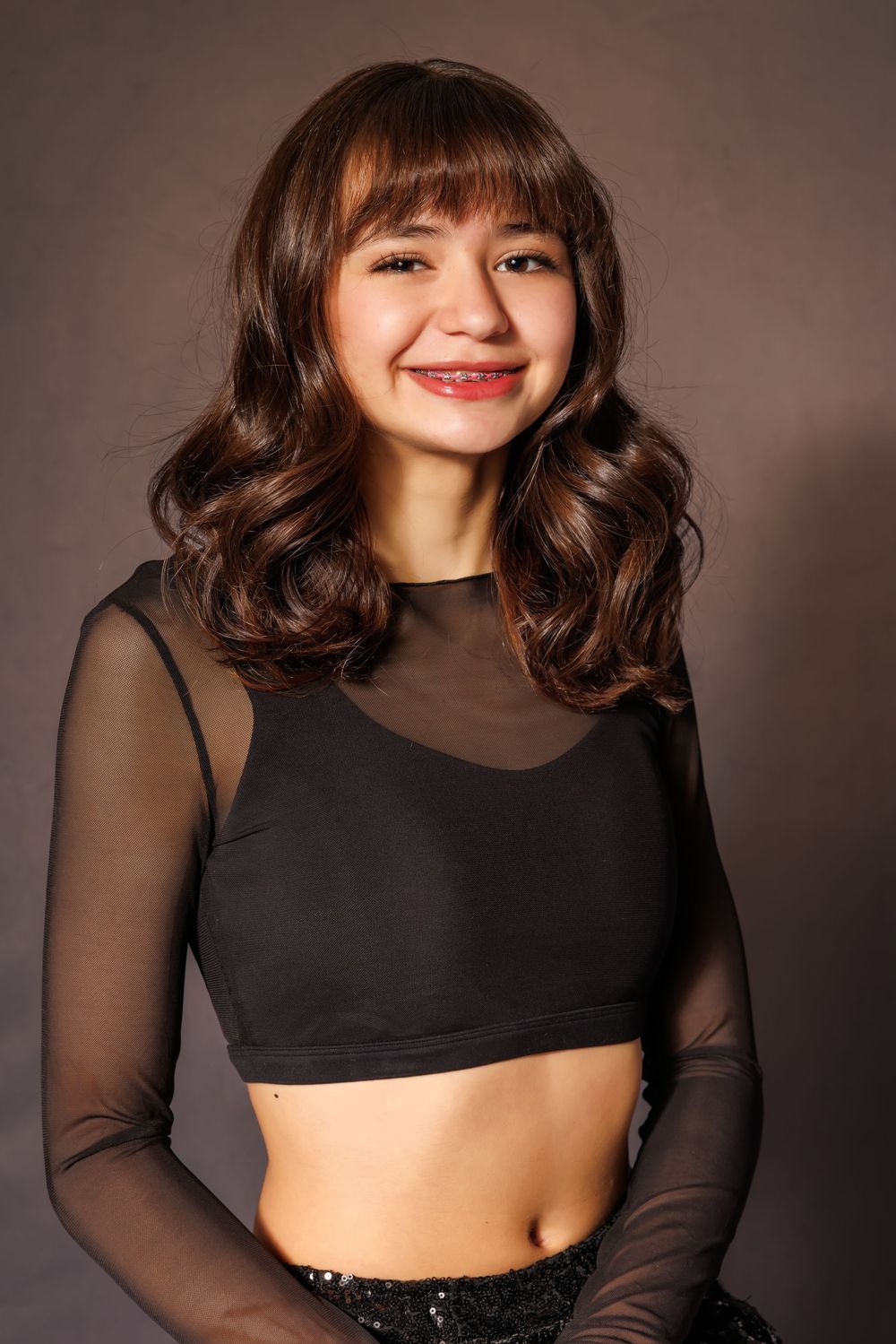 Teenage girl with curly brown hair smiling in black mesh crop top against dark studio background.