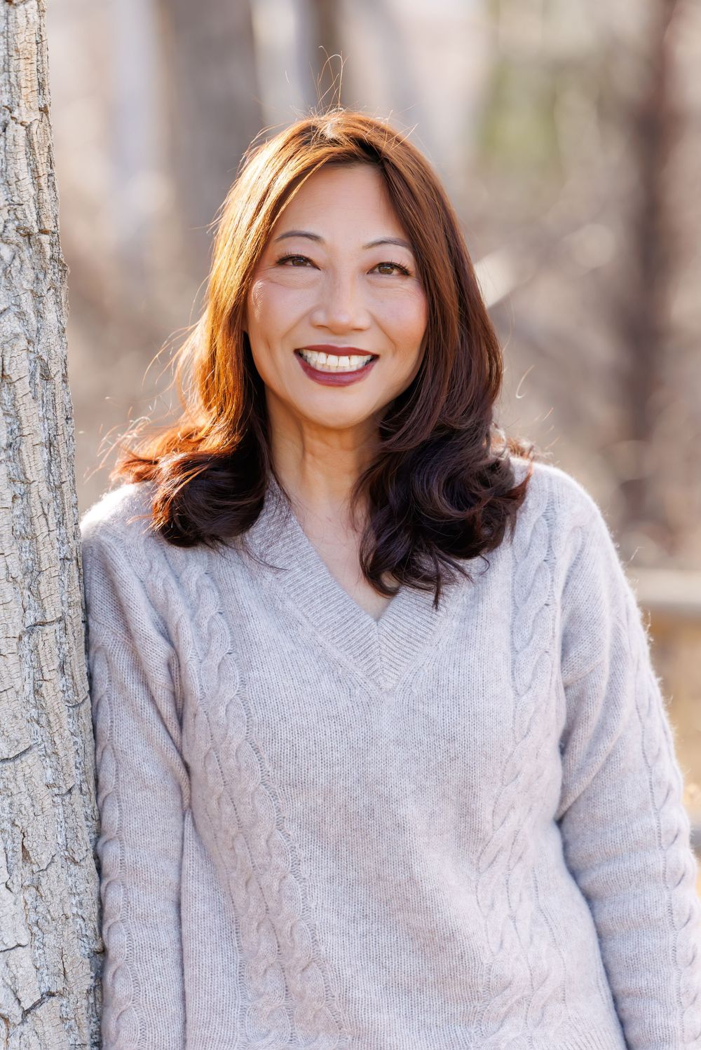 A person wearing a light gray sweater poses outdoors against a blurred winter forest background.