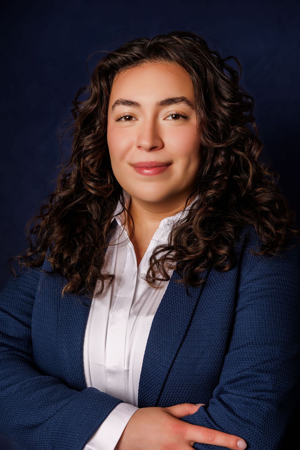 Professional woman with curly brown hair in blue blazer and white shirt, arms crossed, posing for corporate headshot.