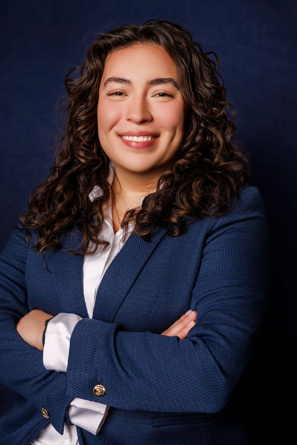 Professional woman in blue blazer with curly hair smiling confidently with arms crossed against dark background.