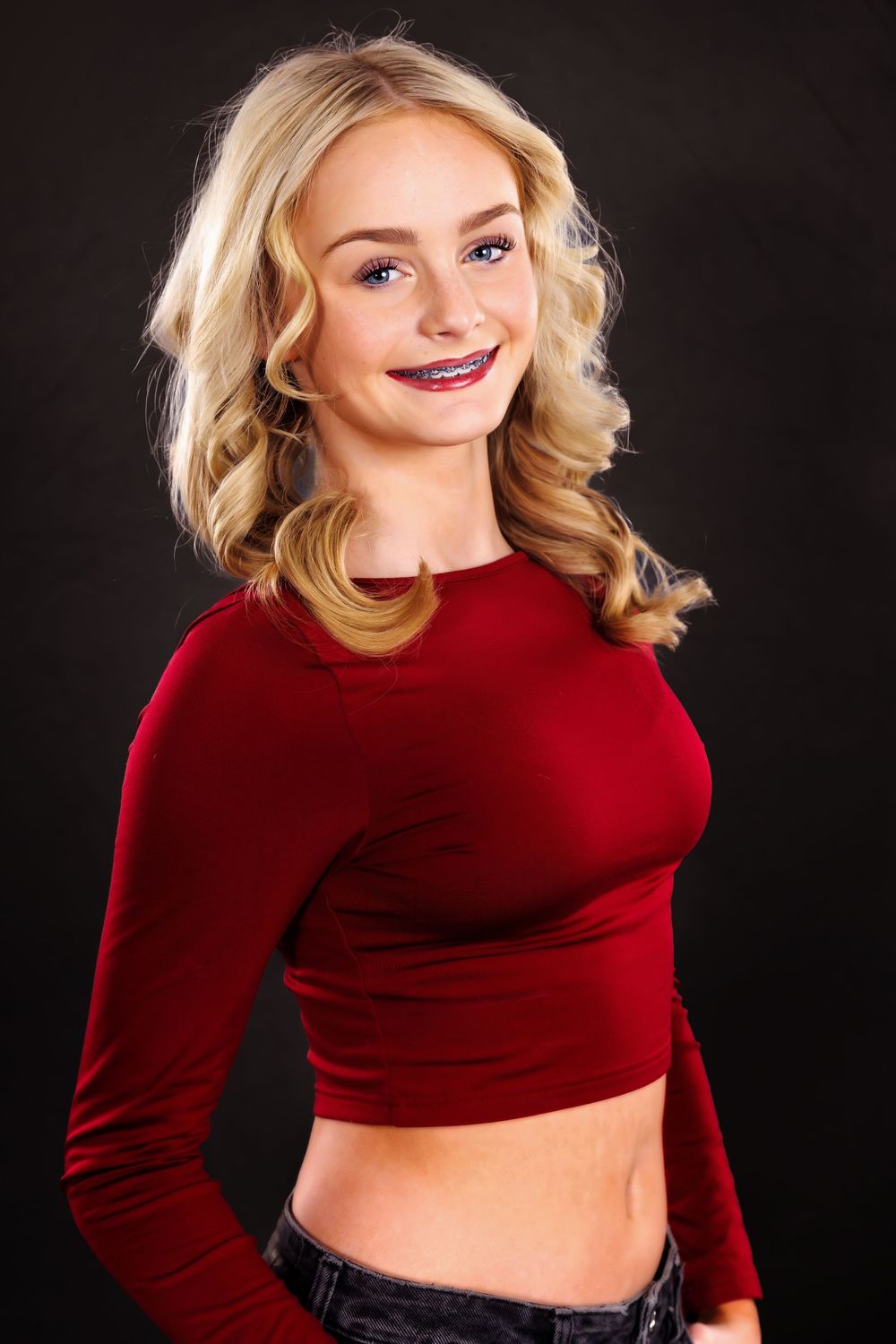 Smiling blonde teen girl with braces wearing a red crop top posing against a dark background.