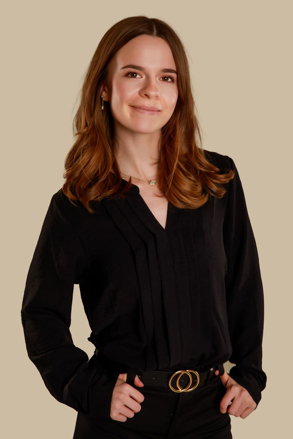 Professional headshot with brown wavy hair wearing black top and designer belt against beige background.