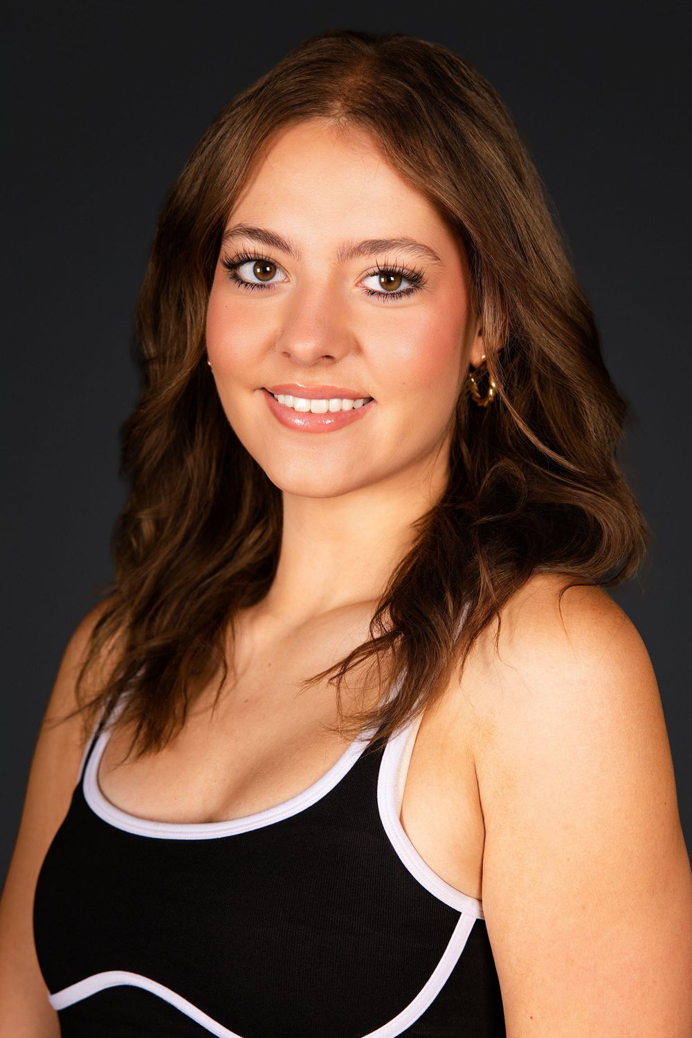 Young woman with brown wavy hair smiling, wearing a black tank top with white trim against a dark background.