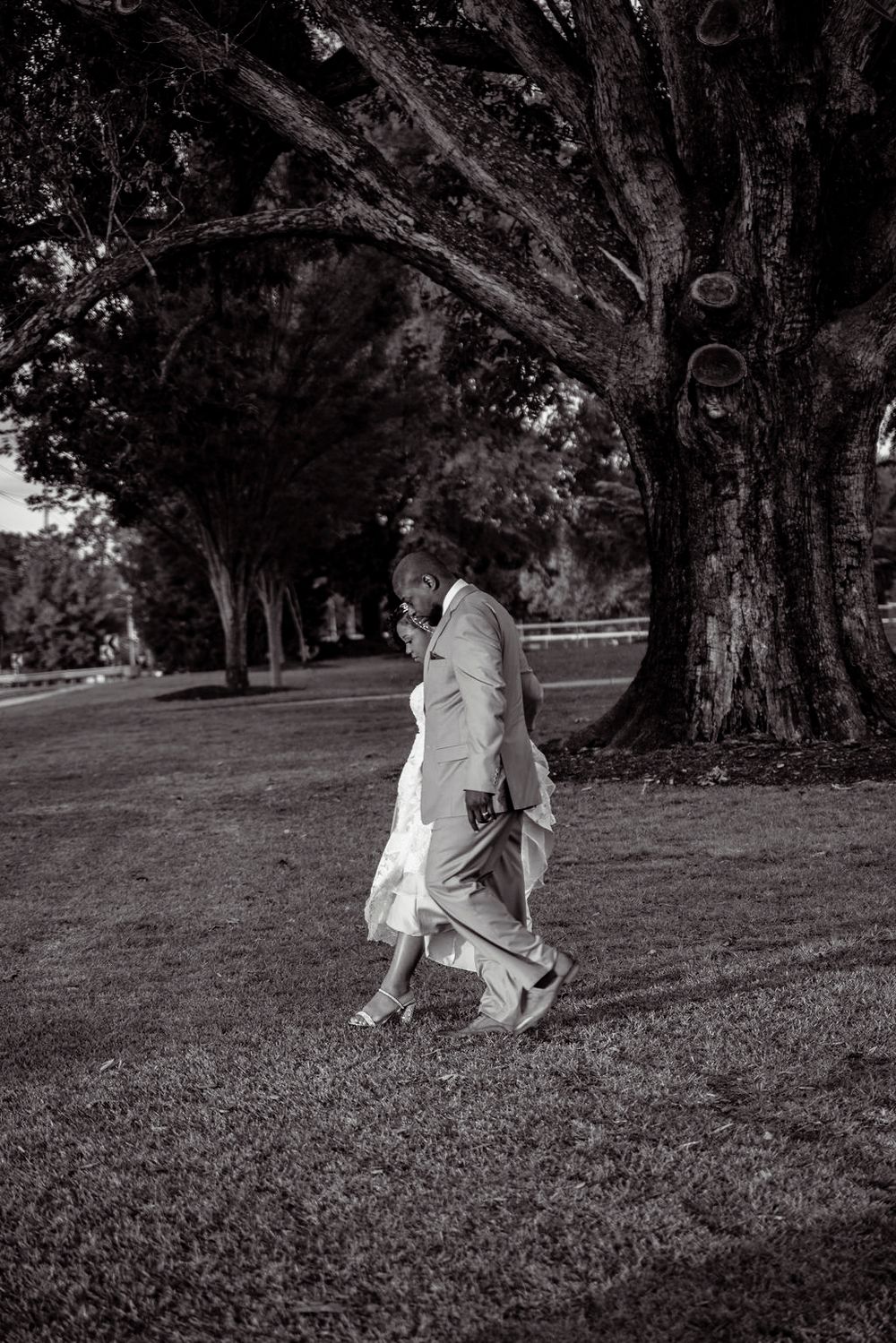 A person in white formal attire walks along a tree-lined path in black and white photography.