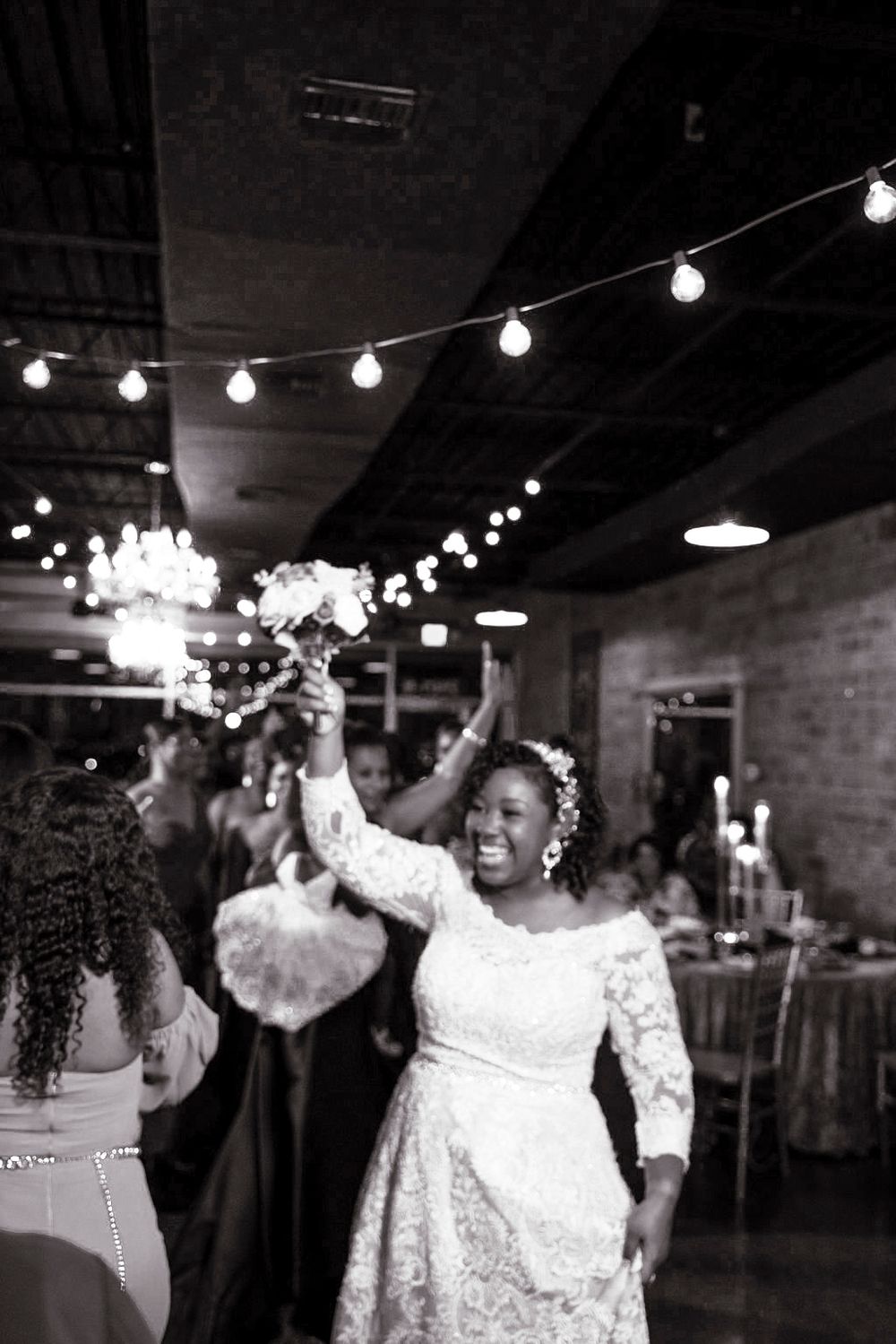 A joyful person in a white lace dress celebrates at a wedding reception under string lights and chandeliers.
