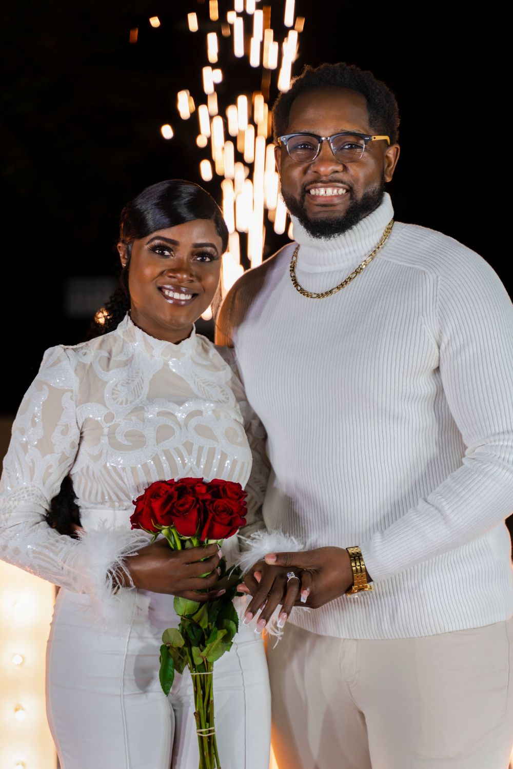 A joyful couple in matching white outfits poses with red roses against a backdrop of sparkling lights.