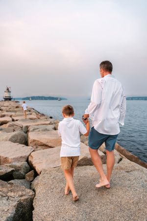 Barefoot figures walk along a rocky coastal shoreline during sunset.