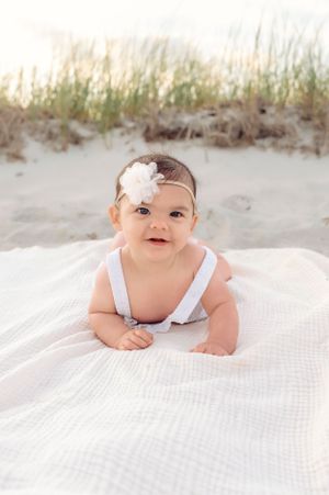 Baby in white outfit lying on blanket with beach grass in background.