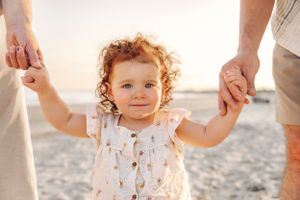 Child with curly hair wearing white patterned outfit on beach at sunset.