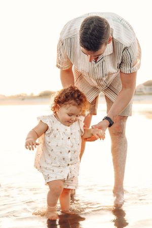 Parent and child exploring shallow water together on beach at sunset.