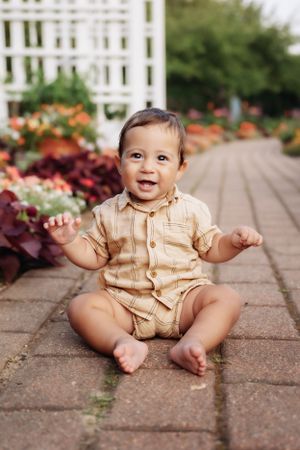 Baby in tan romper sitting on brick pathway by white fence and flowers.
