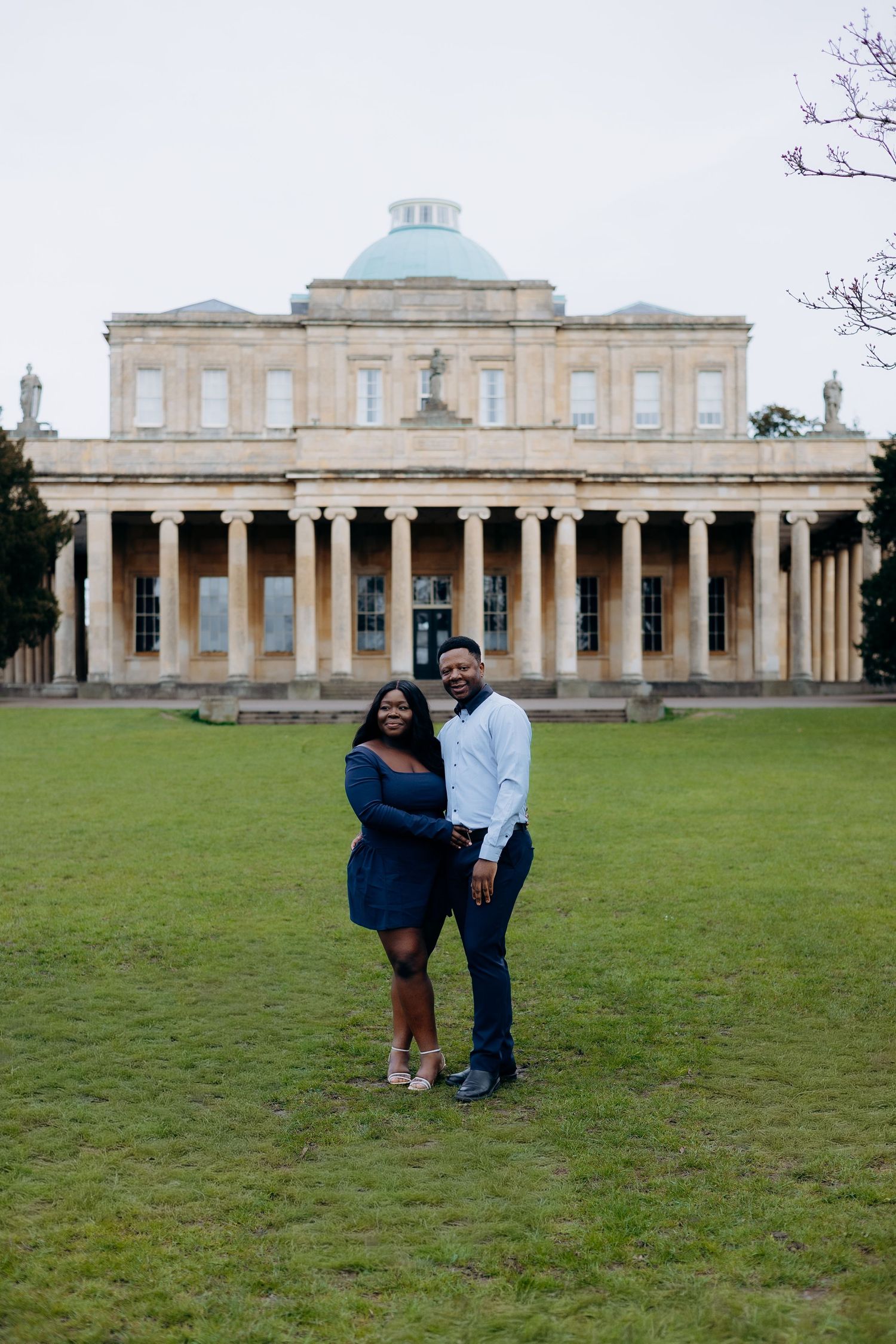 Couple smiling and embracing on green lawn in front of a grand neoclassical building with columns and blue dome in Cheltenham.