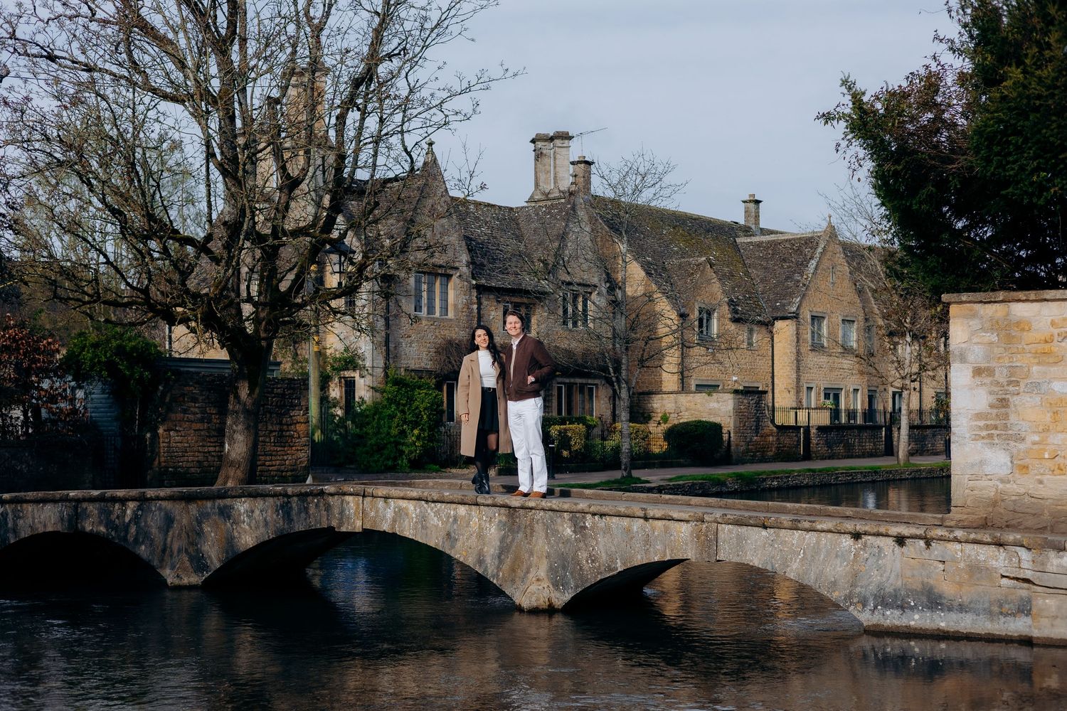 Couple standing on a stone bridge over a calm river in the Cotswolds village of Bourton-on-the-Water.