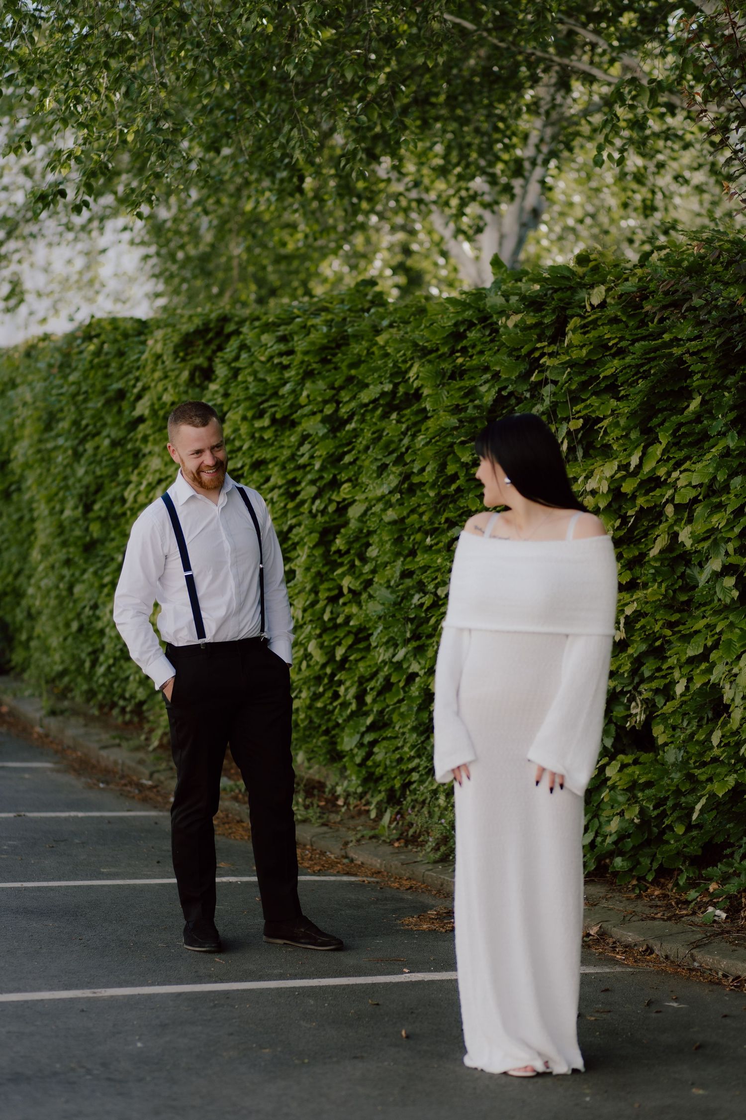A couple stand facing each other on a paved path with green foliage in the background for their pre wedding photoshoot in Cheltenham.