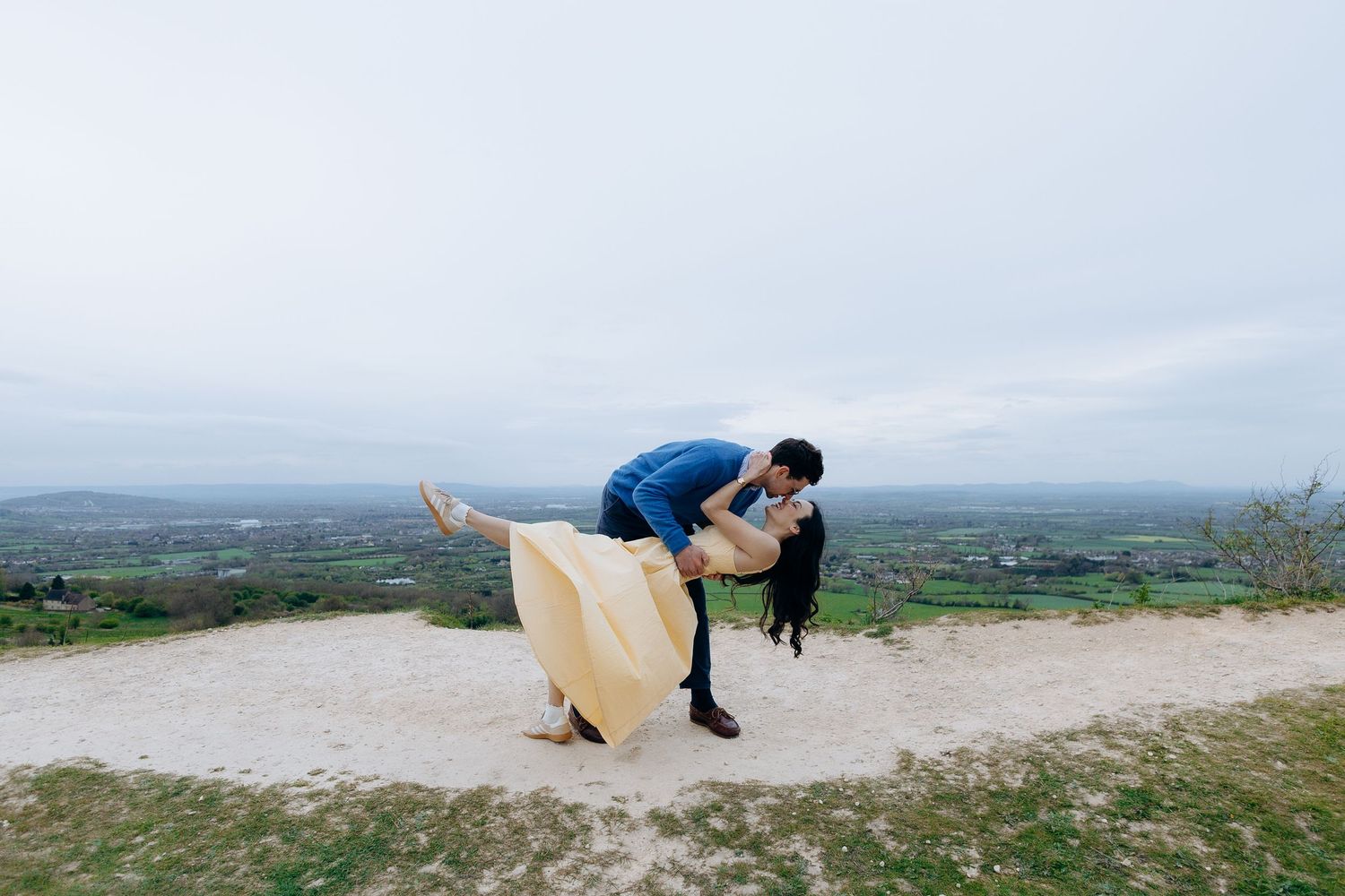 Couple sharing a romantic dip kiss on a Gloucestershire hilltop with scenic green countryside views in the background for their engagement.