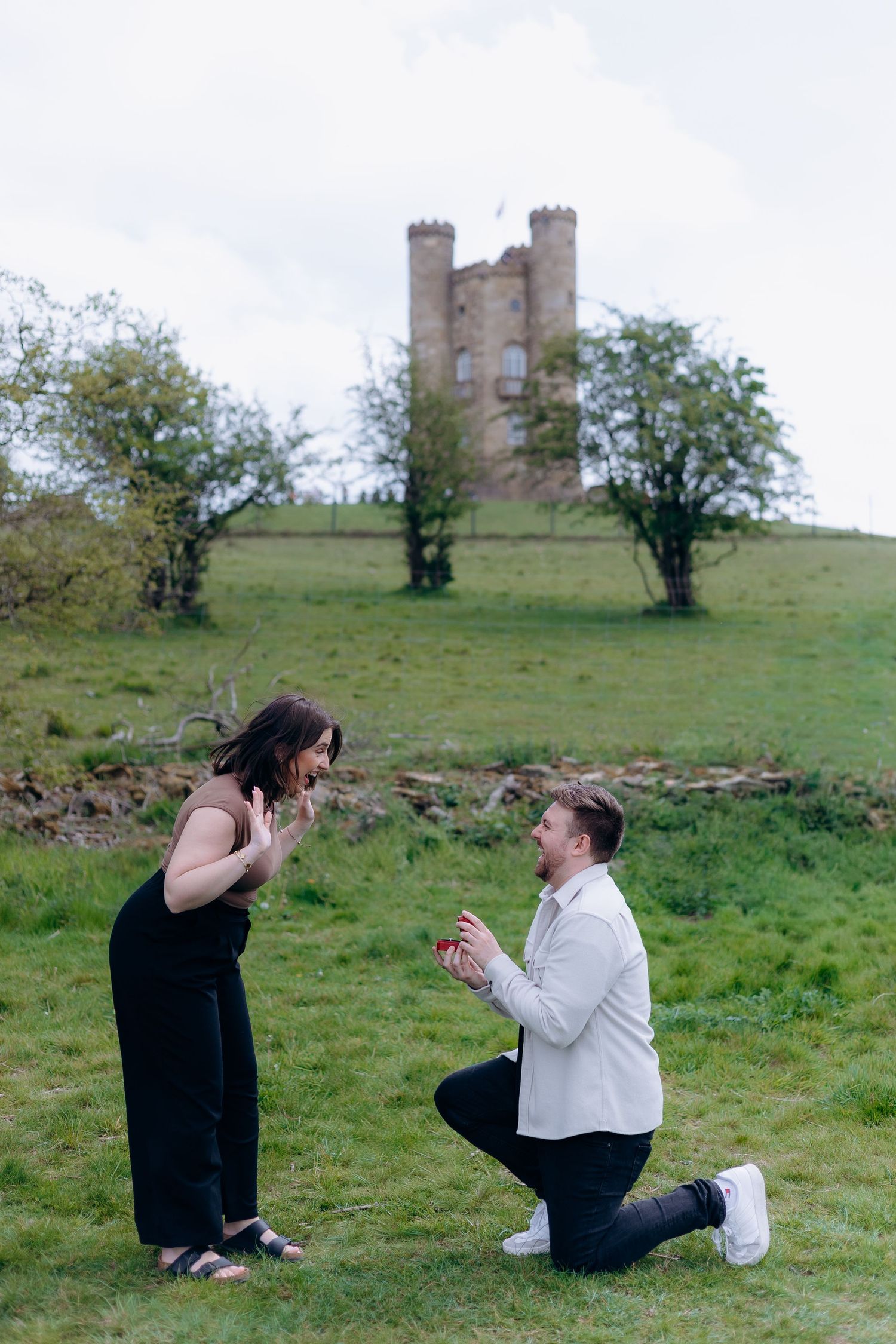 Man proposing on one knee to woman outdoors with Broadway tower on hill behind them.