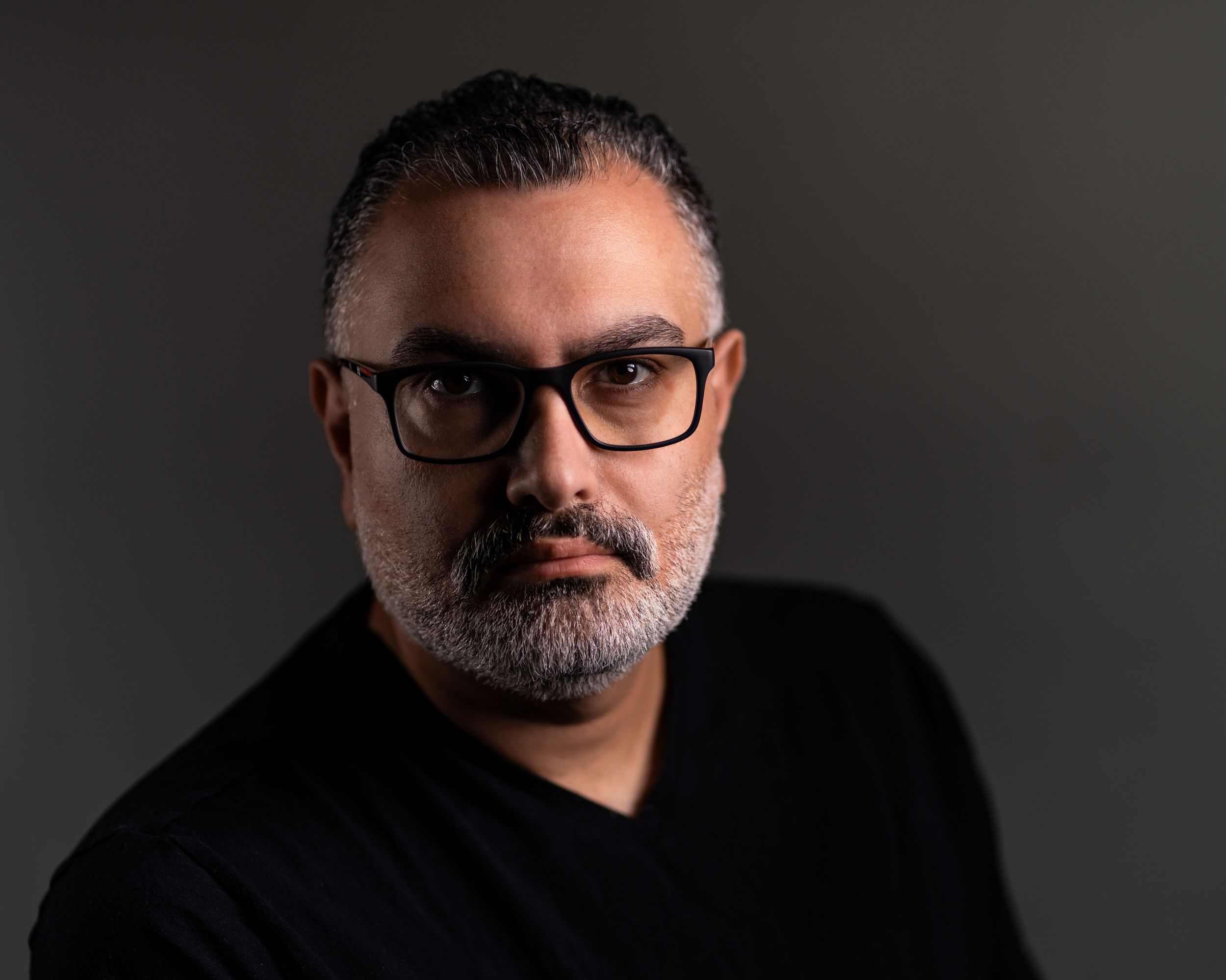 Dramatic portrait of a bearded man with glasses wearing a black shirt against dark background.
