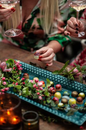Hands holding champagne glasses over a decorated platter with colorful berries and flowers.