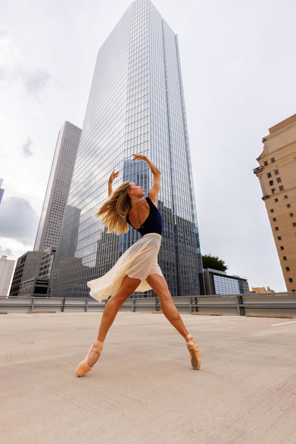Ballet dancer poses gracefully against modern glass skyscraper in urban setting.