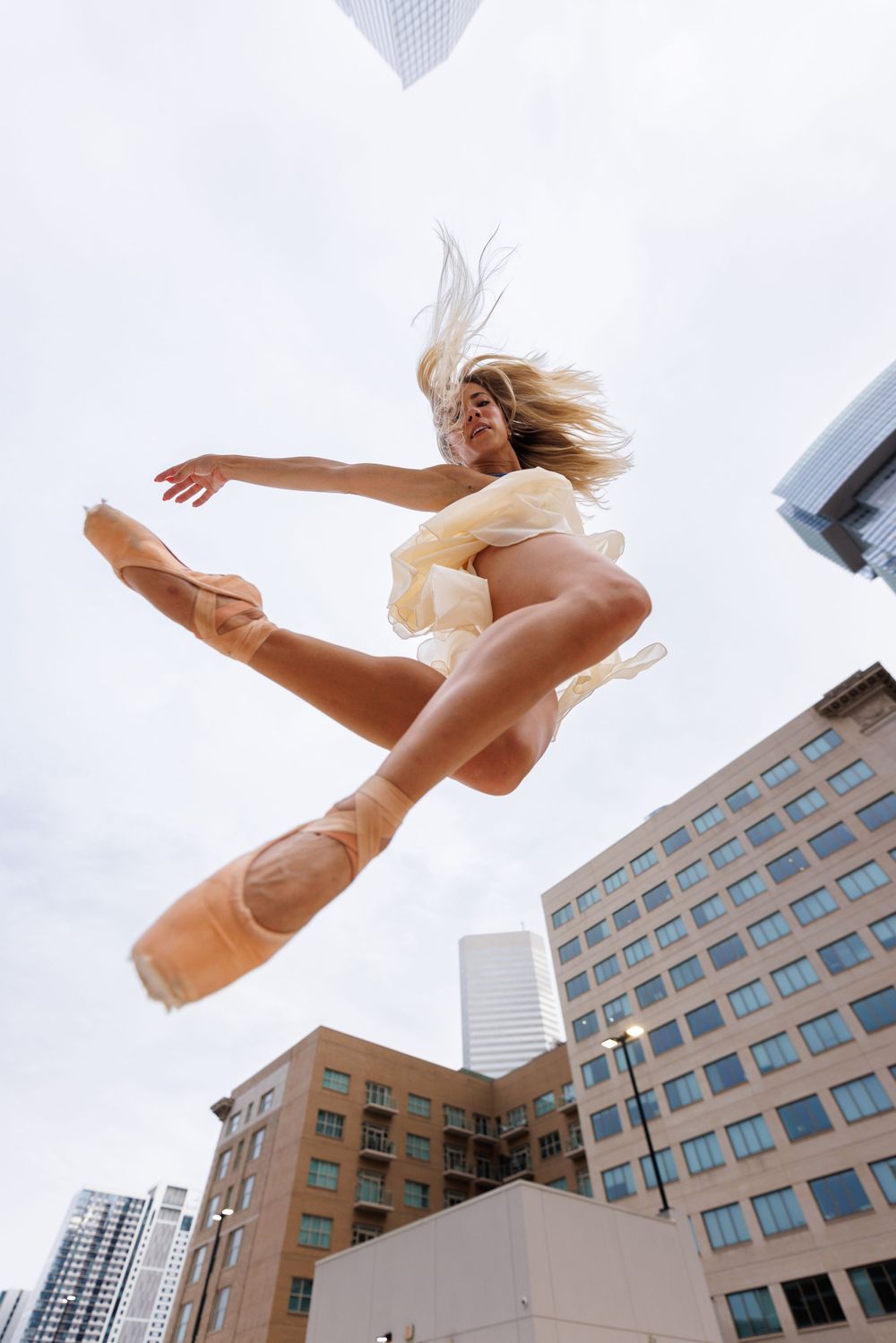A ballet dancer in a flowing dress leaps gracefully between urban buildings against a bright sky.