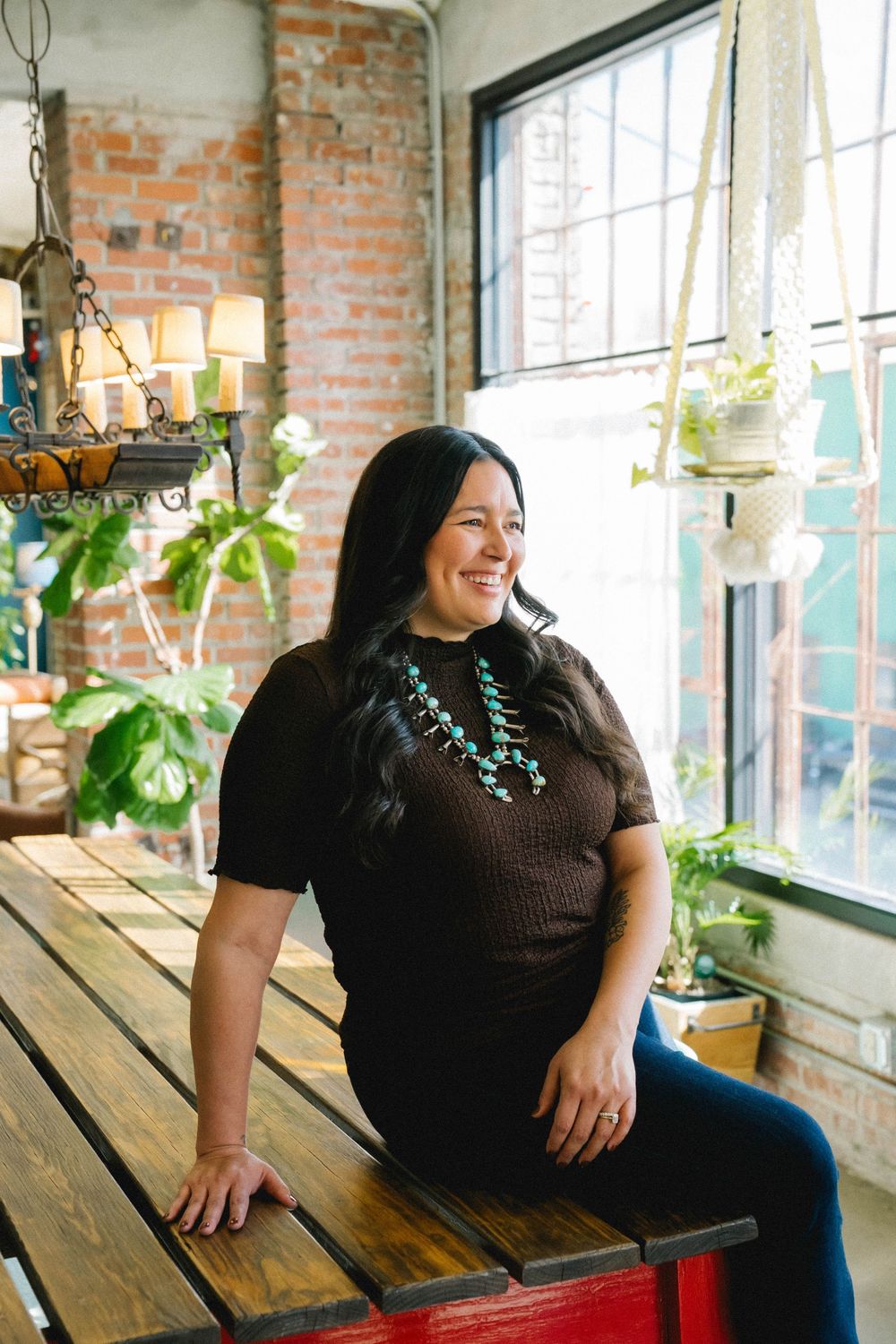 A smiling woman in a brown top and turquoise necklace sits by a window in a rustic brick-walled cafe with hanging plants.