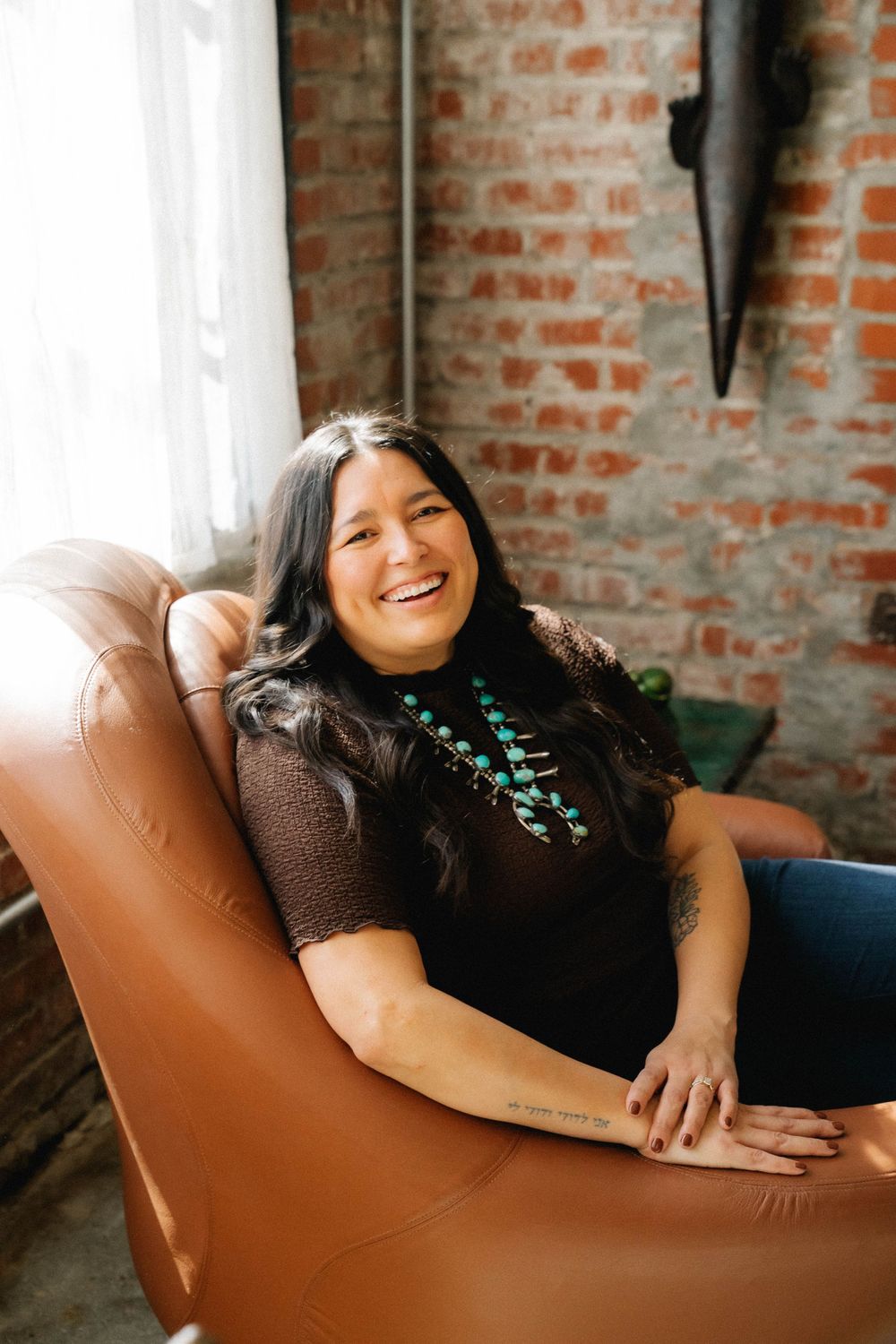 A smiling woman with long dark hair sits in a brown leather chair, wearing a turquoise necklace and dark top near a brick wall.